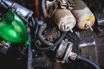 An engine compartment featuring various components, including two fuel filters attached to hoses, an electrical connection, and a funnel-like green plastic bottle. The filters are dusty and rest on a dark, greasy surface, possibly indicating a used or older engine.