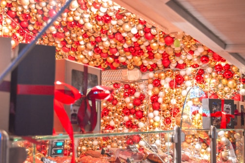 A festive display is adorned with an abundance of red, gold, and white ornaments hanging from the ceiling and walls. Gift boxes wrapped with red ribbons are placed on a glass counter that holds various items. Twinkling lights enhance the joyful atmosphere, and reflections create a warm and inviting scene.