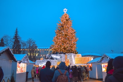 A festive scene at a Christmas market features a large, brightly lit Christmas tree with a star on top. Surrounding the tree are small market stalls decorated with lights and snow. A crowd of people, bundled up in winter clothing, gather around the stalls and tree, enjoying the holiday atmosphere.