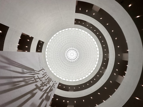 A geometric and symmetrical view of a dome from the inside, showcasing a circular pattern with a central light source. The structure consists of multiple layers of concentric shapes with intricate detailing. The space is illuminated by evenly spaced lights along the circular frames, creating a harmonious and spacious atmosphere. Shadows of text are visible on a large, smooth surface.
