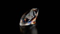 A polished and faceted gemstone, likely a diamond, stands out against a deep black background. The gem's surface reflects and refracts light, creating a spectrum of colors including blues, oranges, and whites across its facets. The overall shape is elongated with precise edges and sharp corners.