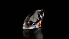 A polished and faceted gemstone, likely a diamond, stands out against a deep black background. The gem's surface reflects and refracts light, creating a spectrum of colors including blues, oranges, and whites across its facets. The overall shape is elongated with precise edges and sharp corners.