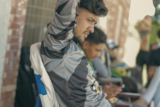 A young man wearing a sports jersey leans forward, appearing focused and thoughtful. He is seated outdoors, possibly on a bench near a brick wall. Behind him, another person is visible but out of focus, holding a mobile device.