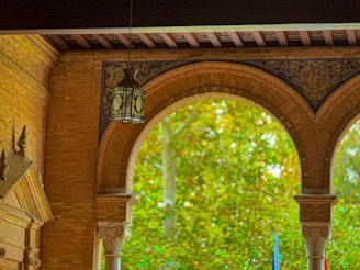 A brick archway adorned with intricate designs frames a view of lush green foliage outside. A vintage lantern hangs from the ceiling, casting a calm and elegant atmosphere. Architectural details like columns and decorative patterns enhance the aesthetic appeal.