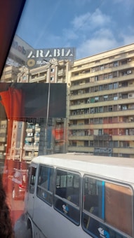 A bustling urban street scene captures several multi-story buildings with a large 'Arabia' sign on one of the rooftops. The image includes a bus and a car on the road, visible through a bus window with red reflections.