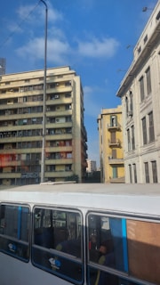 Urban scene featuring tall buildings with multiple windows and balconies, a bus visible in the foreground with reflections on its windows, and a clear blue sky overhead. The buildings exhibit signs of wear with air conditioning units attached and some balconies. A streetlight and electric wires are also present.