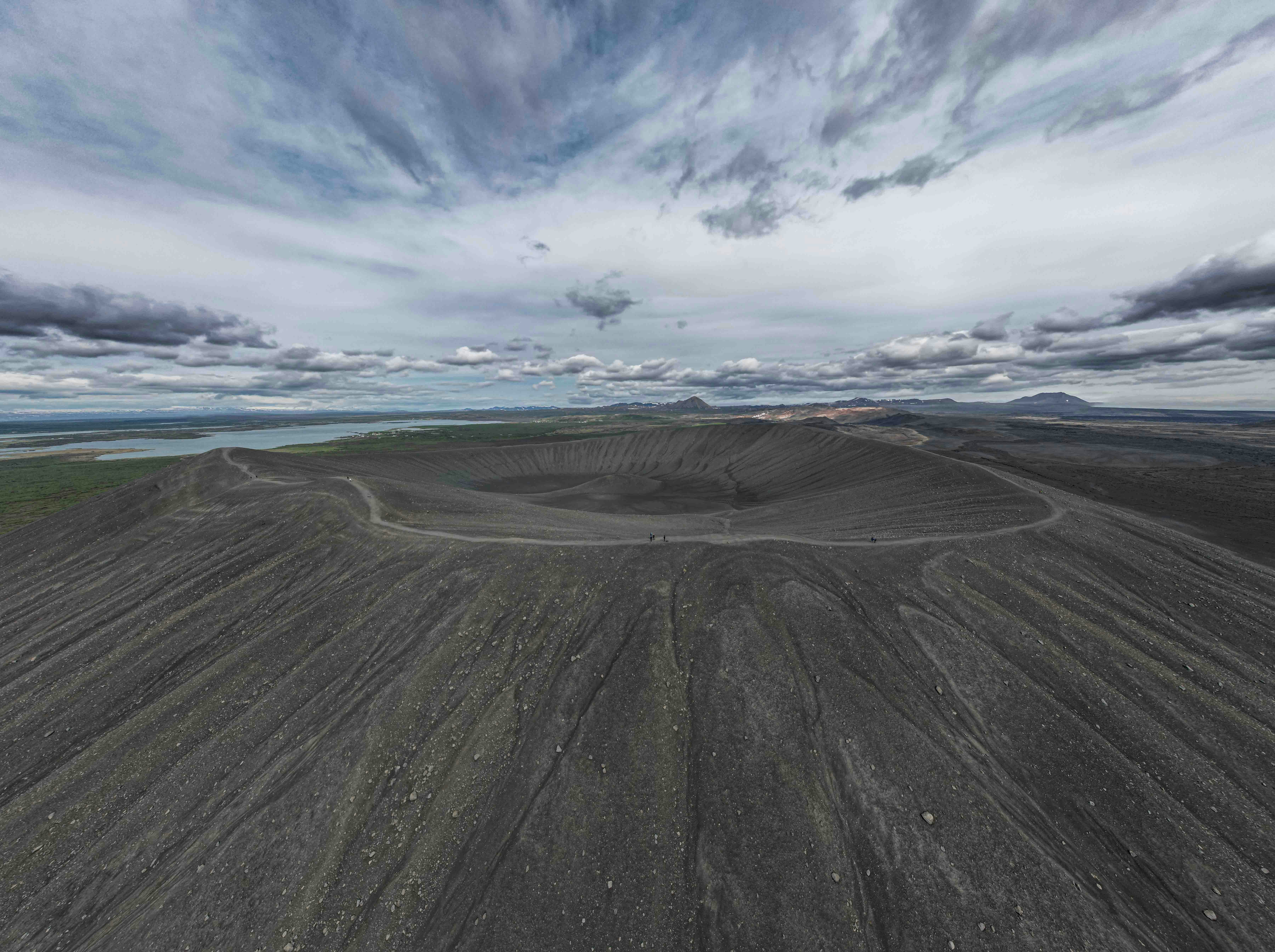 an aerial view of a dirt field with clouds in the sky, A panoramic view of a vast, empty volcanic crater with steep, textured sides under a dynamic sky with scattered clouds. The horizon shows a distant view of a calm body of water and a hint of greenery.
