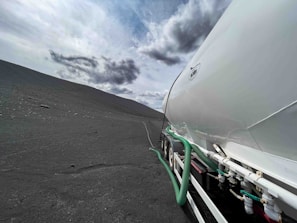 A large tanker vehicle is situated on a barren, rocky landscape under a partly cloudy sky. The tanker appears white with green hoses and is parked on an expansive terrain that is mostly devoid of vegetation or other structures.