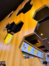 A modern interior with hexagonal cutouts in a wooden wall, revealing digital menu screens displaying various food and drink items. The area is well-lit with a warm ambiance, featuring sleek, dark metallic accents around the cutouts.