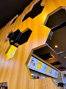 A modern interior with hexagonal cutouts in a wooden wall, revealing digital menu screens displaying various food and drink items. The area is well-lit with a warm ambiance, featuring sleek, dark metallic accents around the cutouts.