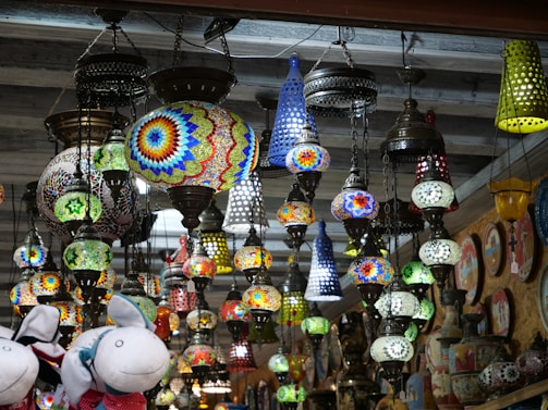 A variety of vibrant, intricately designed hanging lamps with mosaic patterns are displayed. The lamps are suspended from the ceiling with metal chains, creating an eclectic and colorful array. In the forefront, plush toy animals are visible.