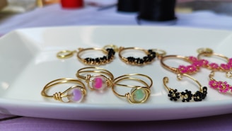 A collection of gold wire rings displayed on a white rectangular dish. Some rings feature colorful beads, including pastel pink, green, and black, with designs appearing delicate and ornamental.