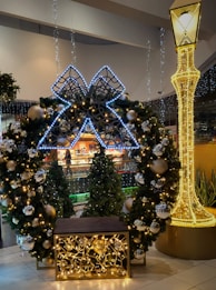 A festive holiday display featuring a large decorative wreath adorned with glowing lights and shiny ornaments. The wreath is complemented by a large illuminated bow positioned at the top. Nearby, a tall light-up structure resembling a street lamp adds to the festive ambiance, and a row of decorated Christmas trees in the background enhances the holiday theme.