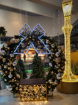 A festive holiday display featuring a large decorative wreath adorned with glowing lights and shiny ornaments. The wreath is complemented by a large illuminated bow positioned at the top. Nearby, a tall light-up structure resembling a street lamp adds to the festive ambiance, and a row of decorated Christmas trees in the background enhances the holiday theme.