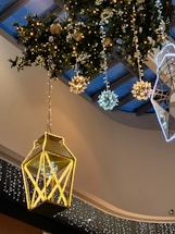 A festive indoor decoration featuring a large, illuminated lantern suspended from the ceiling. Surrounding it are several spherical ornaments and decorative snowflakes, all elegantly lit, hanging from greenery with additional small lights. The ceiling has a contemporary design with glass panels.