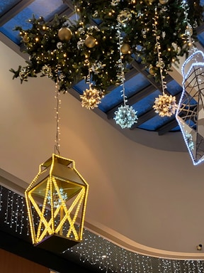 A festive indoor decoration featuring a large, illuminated lantern suspended from the ceiling. Surrounding it are several spherical ornaments and decorative snowflakes, all elegantly lit, hanging from greenery with additional small lights. The ceiling has a contemporary design with glass panels.