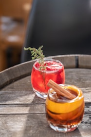 Two cocktails are positioned on a wooden table. The front cocktail is garnished with a cinnamon stick and an orange peel, showing a rich amber color. The cocktail in the background has a bright red hue and is garnished with a sprig of fresh thyme.