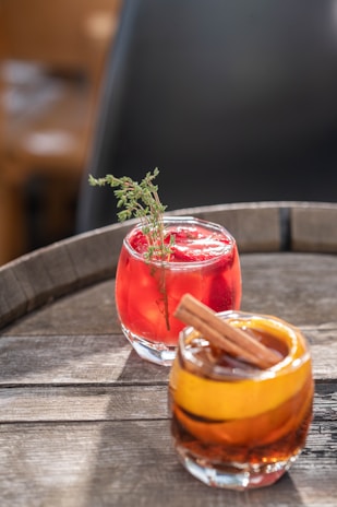 Two cocktails are positioned on a wooden table. The front cocktail is garnished with a cinnamon stick and an orange peel, showing a rich amber color. The cocktail in the background has a bright red hue and is garnished with a sprig of fresh thyme.