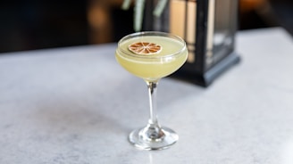 A cocktail in a coupe glass garnished with a dried citrus slice sits on a light gray surface. A lantern is softly blurred in the background, adding a touch of elegance.
