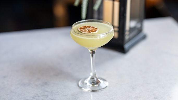 A cocktail in a coupe glass garnished with a dried citrus slice sits on a light gray surface. A lantern is softly blurred in the background, adding a touch of elegance.
