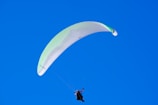 A paraglider is soaring through a clear blue sky. The parachute wing is primarily white with subtle green accents. A person is suspended below, wearing a harness and gear.