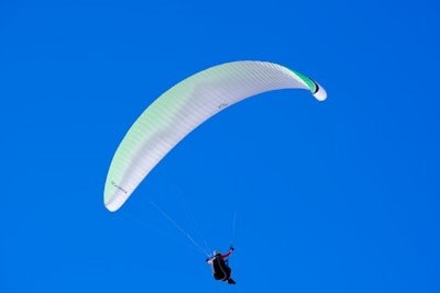 A paraglider is soaring through a clear blue sky. The parachute wing is primarily white with subtle green accents. A person is suspended below, wearing a harness and gear.