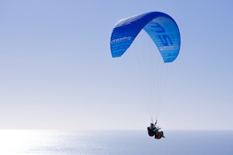 A paraglider is soaring high in the sky over the ocean, with a vibrant blue parachute. The sun reflects off the water, creating a shimmering effect. The scene conveys a sense of freedom and adventure.