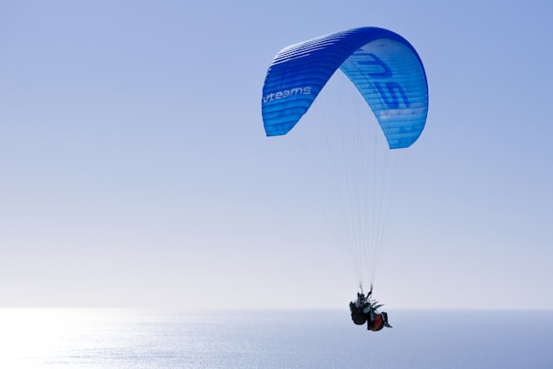 A paraglider is soaring high in the sky over the ocean, with a vibrant blue parachute. The sun reflects off the water, creating a shimmering effect. The scene conveys a sense of freedom and adventure.