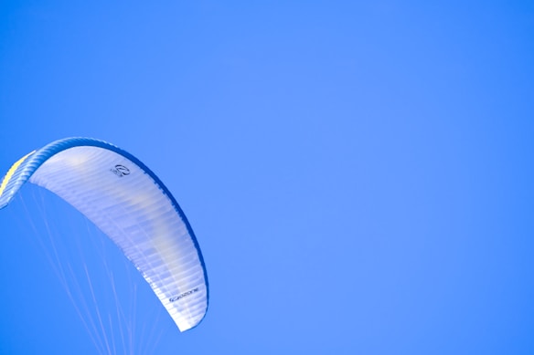 A gliding parachute with a light-colored, semi-transparent fabric against a bright blue sky. The parachute displays sleek, curved lines and subtle design details, showing gentle motion and openness.