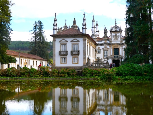 A grand historic mansion with ornate architectural features, including intricate columns and decorative statues, is situated amidst lush greenery. The building is reflected in a tranquil body of water in the foreground, and a row of trees stands majestically in the background.