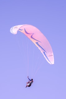 A person is paragliding against a clear sky. The paraglider is colored with shades of pink and blue, creating a serene scene as the person is suspended in the air with a sense of freedom and adventure.