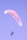 A person is paragliding against a clear sky. The paraglider is colored with shades of pink and blue, creating a serene scene as the person is suspended in the air with a sense of freedom and adventure.