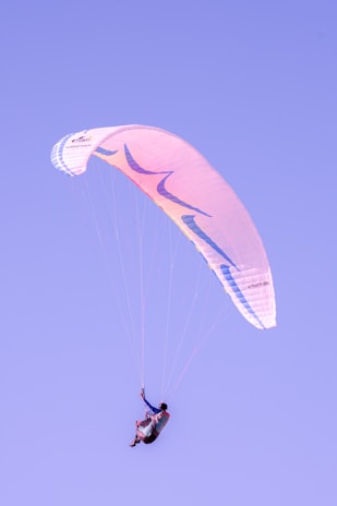 A person is paragliding against a clear sky. The paraglider is colored with shades of pink and blue, creating a serene scene as the person is suspended in the air with a sense of freedom and adventure.