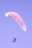 A person is paragliding against a clear sky. The paraglider is colored with shades of pink and blue, creating a serene scene as the person is suspended in the air with a sense of freedom and adventure.