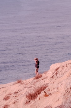 A person stands on a rocky, sandy cliff overlooking a vast body of water. The individual appears to be looking through binoculars, with one hand shielding their eyes from the sunlight. The scene conveys a sense of tranquility and exploration.
