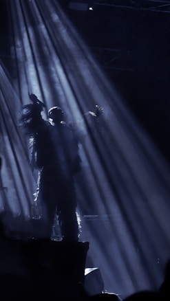 A silhouetted figure stands on a stage, illuminated by dramatic beams of light that create a mysterious and atmospheric effect. The scene is dark, with faint outlines and textures suggesting movement or dynamic action.