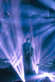 A performer is standing on stage surrounded by beams of light. The lighting creates dramatic effects, casting shadows and highlights. The performer is wearing a textured outfit, and the stage has instruments arranged around.
