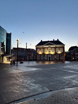 A town square during early evening or dusk with a prominently lit classical building at the center. The building has 'Sneider Zijlstra' written on it. Birds are seen flying in the sky above. There's a modern glass building on the left, street lamps provide illumination, and the ground looks wet, likely from recent rain. A few people and vehicles are visible.