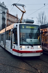 A modern white tram decorated with festive lights travels along cobblestone tracks in an urban setting, against a backdrop of historic buildings. The tram displays route number 5 and destination 'Stefanikova Ct'. Overhead electric wires are visible, and the surrounding area appears overcast.