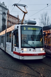 A modern white tram decorated with festive lights travels along cobblestone tracks in an urban setting, against a backdrop of historic buildings. The tram displays route number 5 and destination 'Stefanikova Ct'. Overhead electric wires are visible, and the surrounding area appears overcast.