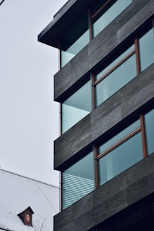 A modern building with large glass windows and a flat roof, featuring dark gray paneling. Adjacent to the structure is a traditional house with a pitched roof and a small circular attic window, creating a contrast between contemporary and classic architectural styles.