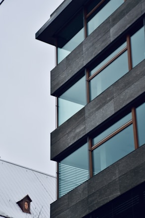 A modern building with large glass windows and a flat roof, featuring dark gray paneling. Adjacent to the structure is a traditional house with a pitched roof and a small circular attic window, creating a contrast between contemporary and classic architectural styles.