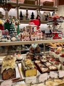 A festive display of Christmas-themed decorations and an assortment of desserts. The scene includes a gingerbread train adorned with candy canes, a carousel, decorated Christmas trees, and small houses. Various desserts such as fruit-topped cakes, slices of chocolate brownie, and layered mousse cups are placed on the countertop. Holiday decorations and colorful treats create a cheerful and celebratory atmosphere.