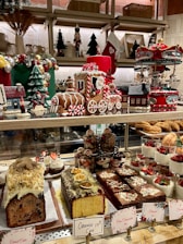 A festive display of Christmas-themed decorations and an assortment of desserts. The scene includes a gingerbread train adorned with candy canes, a carousel, decorated Christmas trees, and small houses. Various desserts such as fruit-topped cakes, slices of chocolate brownie, and layered mousse cups are placed on the countertop. Holiday decorations and colorful treats create a cheerful and celebratory atmosphere.