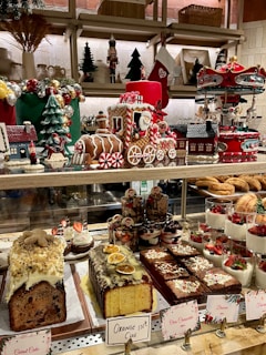 A festive display of Christmas-themed decorations and an assortment of desserts. The scene includes a gingerbread train adorned with candy canes, a carousel, decorated Christmas trees, and small houses. Various desserts such as fruit-topped cakes, slices of chocolate brownie, and layered mousse cups are placed on the countertop. Holiday decorations and colorful treats create a cheerful and celebratory atmosphere.