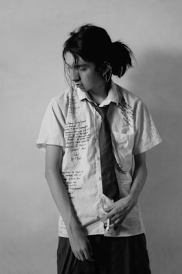 A person with tousled hair wearing a white shirt covered in handwritten text and a loosely tied necktie. The person is looking down and has a thoughtful expression. The image is in black and white, giving it a classic and introspective feel.