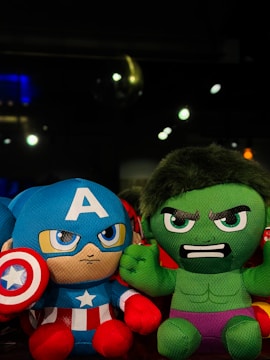 Two plush toys resembling superhero characters with one in blue and red, holding a shield, and another in green with an angry expression. They are displayed against a dark background with some blurred lights.