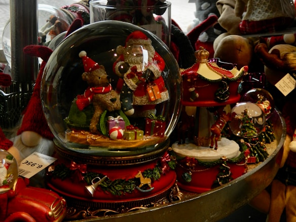 A collection of festive snow globes featuring holiday-themed figures such as Santa Claus and reindeer, adorned with vibrant decorations including gifts and a small carousel. The globes are placed on a display table surrounded by other seasonal ornaments and plush toys, enhancing the holiday atmosphere.