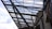 Steel beams and rods form a complex, geometric lattice structure above a modern architectural building. The sky visible in the background is partly cloudy, providing contrast to the industrial elements.
