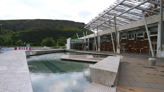 A modern architectural structure with a water feature in the foreground. The building has a unique design with an overhanging metal framework and large glass windows. The background features a lush green hillside, adding a natural contrast to the urban setting.